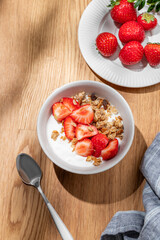 Granola or muesli with natural greek yogurt, strawberry in a bowl on a wooden table with berries and shadows.
