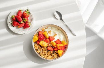 Granola or muesli with natural greek yogurt, strawberry and peach in a bowl on a white table near window