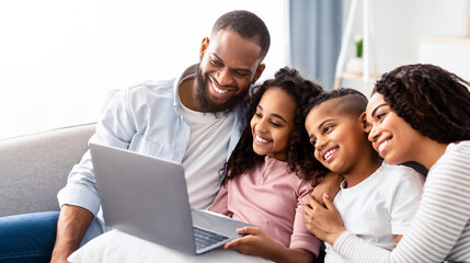 Portrait of cheerful African American parents and their happy children using laptop on pillow, watching movie or having online video conference, sitting on the couch in living room at home together
