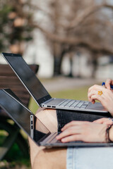Three cheerful students study and work together outdoors on a sunny day, using laptops and notebooks while sitting on a park bench