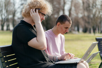 Three cheerful students study and work together outdoors on a sunny day, using laptops and notebooks while sitting on a park bench