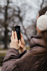 A young woman in a winter coat and earmuffs captures a moment on her smartphone in a city park on a cold day