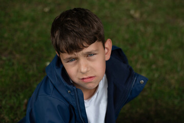 A young boy with a serious look on his face sitting outside on a grassy area in daytime. He wears a blue jacket and white shirt. The boy has indifferent, sad look