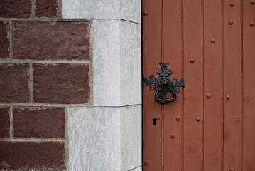 A detailed view of a textured, brown wooden door featuring an ornate black metal knocker against brick and stone wall. Old decorative lock on an old church door
