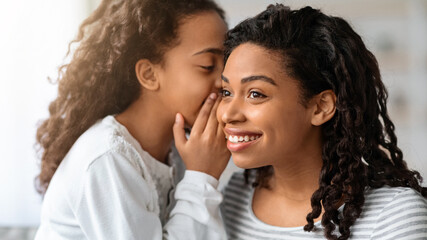 Black girl whispering something to her excited beautiful mother, closeup portrait. African american...