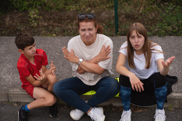 The children are arguing among themselves and the mother asks them to stop and calm down. A family group with mother and two children expressing animated emotions while sitting outdoors