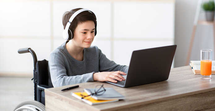 Disabled teen boy listening to music while doing online homework on laptop, indoors. Impaired adolescent using pc to study or communicate remotely, speaking with friends or family on webcam