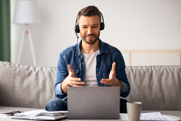 Happy handsome bearded man having video call from home, using headset and laptop while sitting on...