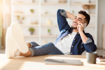 Relaxed arab guy talking on phone while having break, sitting at workdesk with legs up in office or...