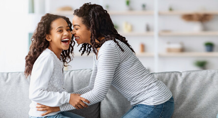Happy african american mom tickling her laughing kid sweet curly girl, sitting together on couch in...