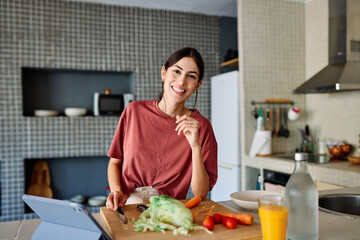 Happy young latina woman having fun preparing food and looking for recipes online using a tabletor...