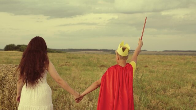 mother son walk through green field sunset. child teenager plays superhero nature. hold woman hand. teamwork. happy family. fancy dress red cloak, crown, sword. woman mother with boy son travels park - Powered by Adobe
