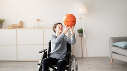 Happy disabled teenager playing with basketball, sitting in wheelchair at home. Paralympic...