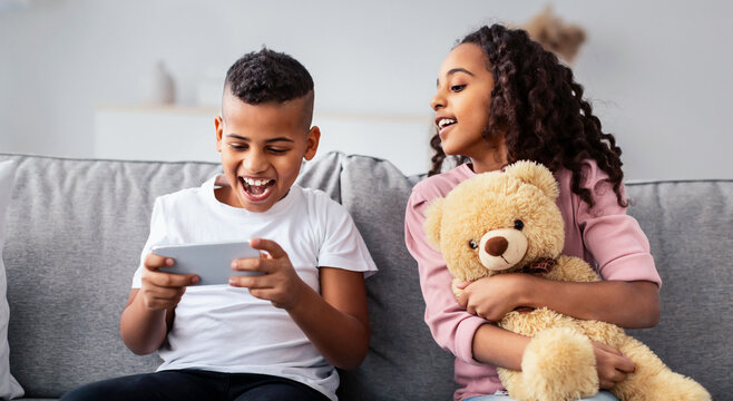 Children Entertainment And Leisure. Portrait of excited black boy playing online mobile game on his smartphone, interested girl looking at the dagdet screen, hugging teddy bear, sitting on couch