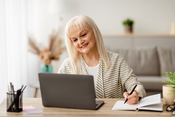 Self Education At Home. Smiling mature woman writing in notebook and using laptop in living room,...