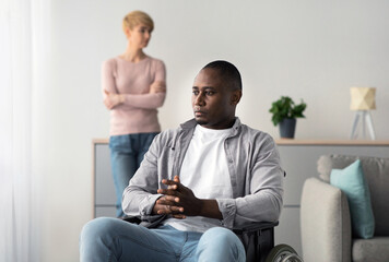 Fototapeta premium Quarrels and problems on self-isolation, covid-19 and after injury. Sad adult african american guy disabled in wheelchair looks at window in living room interior. Upset lady stands aside, silent