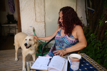 A joyful woman sits on her patio, enjoying coffee, petting her dog, and reviewing notes from her...