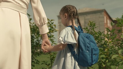 Fototapeta premium mom leads her little daughter with a backpack to school, escort child to preschool education, walk home through schoolyard, first grade student, happy family, kid with school bag by hand with mother.