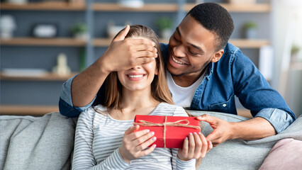 Loving Black Man Covering Eyes And Surprising His White Girlfriend With Romantic Gift At Home, Greeting With Valentine's Day, Birthday Or Anniversary, Mixed Couple Celebrating Holidays Together