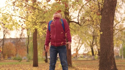 Obraz premium little happy girl with a backpack throws leaves high up and laughs cheerfully, child play in the autumn park, kid walks in the city park, raise her face looking at the golden leaf fall, autumn season.