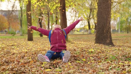 girl with a backpack throws dry autumn leaves up over her head, a school bag on the shoulders of a schoolgirl, a child in a city pack plays with yellow foliage, happy little kidna nature outdoors.