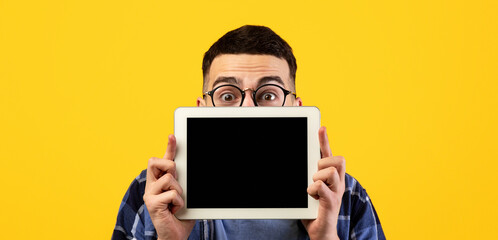 Shocked young guy hiding behind tablet computer with blank screen on orange studio background, mockup for design. Surprised millennial man holding touch pad with space for your app or website