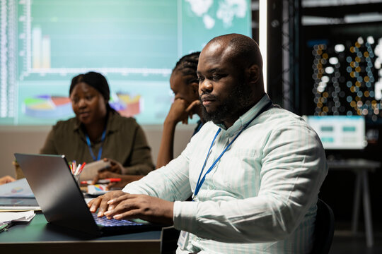 African american male analyst reviewing budget numbers and insight after resource allocation, reviewing data on laptop during night time meeting with his team. After hours night scene.