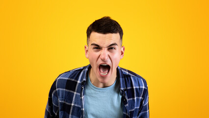 Frustrated young guy shouting in anger, expressing his fury over orange studio background. Enraged...