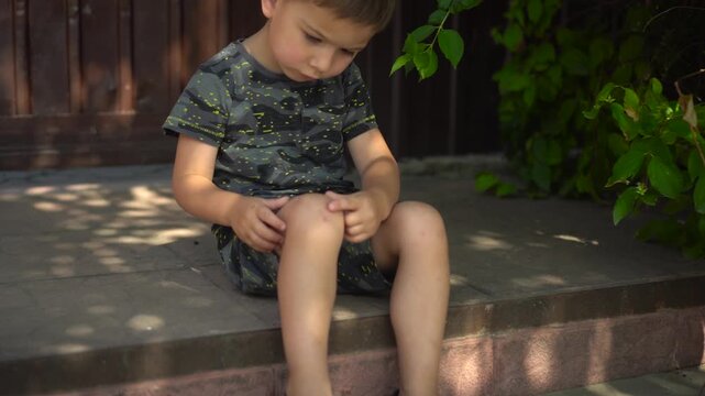 the boy sits on the porch on a hot summer day gently touching a scab on his knee with his finger the camera slowly zooms in toward his face