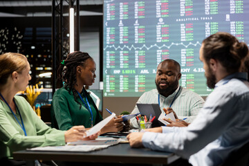 Diverse team of analysts reviewing annual data reports in after hours meeting, developing internal...