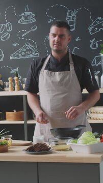 Vertical shot of chef in aron explaining cooking process to his viewers, simultneously tossing vegetables in pan during his kitchen vlog