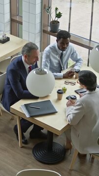 Vertical shot of three intercultural white collar workers sitting by table in cafe and discussing main points of their reports before business conference