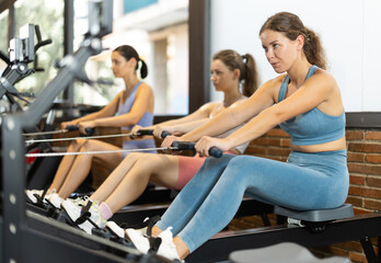 Sportive young girls training on cable row machine in large fitness hall during daytime