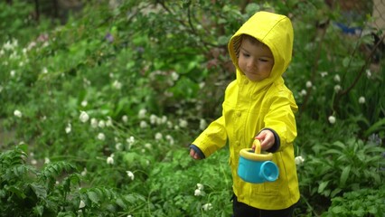 small child in yellow raincoat with hood stands in the rain filling a toy watering can with water against a green garden background