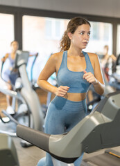 Fototapeta premium Woman in tracksuit at cardio workout, she uses treadmill in gym. European woman doing exercises for body relief