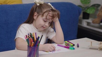 Young girl is sitting at a table with a blue couch behind her. She is drawing with markers and crayons
