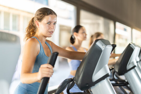 Sportive young woman doing exercises on elliptical trainer in well-equipped gym with large windows