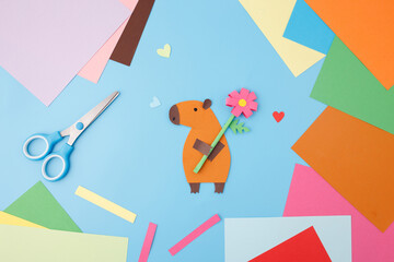 Handmade paper capybara and craft supplies on a blue background. Flat lay composition with scissors, glue stick, colored paper, and cut-out shapes. DIY children's craft and creative hobby concept