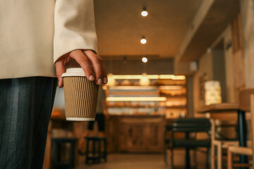 Close-up of hand holding takeaway coffee cup in stylish cafe interior, person wearing elegant jacket and trousers