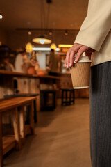 Person holding paper coffee cup inside cozy cafe with wooden tables, wearing elegant jacket and striped trousers