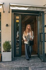 Young woman in beige blazer holding laptop, coffee cup, and shopping bag walking out of modern cafй on sunny day, urban lifestyle concept