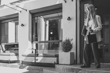 Young woman in beige blazer holding laptop, coffee cup, and shopping bag walking out of modern...