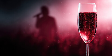 A glass filled with red sparkling liquid is positioned in the foreground, with a blurred figure holding a microphone in the background. Celebration and entertainment
