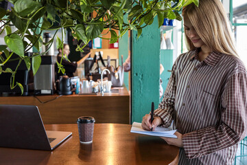 Young woman writing in notebook while smiling at wooden cafe table with laptop and takeaway coffee