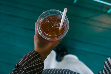 Top view of female hand holding plastic cup with iced tea and straw against green wooden floor