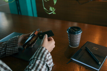 Close-up of person holding bank card and wallet at cafe table with coffee cup, notebook, and pen