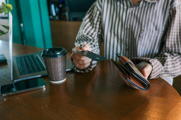 Close-up of woman holding wallet with bank card at cafe table, laptop, smartphone, and coffee cup...