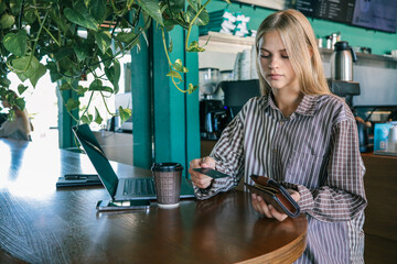 Young woman holding wallet and bank card while working on laptop in cafe, coffee cup and smartphone...