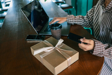 Close-up of hand holding bank card near laptop, coffee cup, and wrapped gift box on cafe table