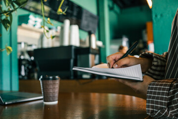 Close-up of hand writing in lined notebook with black pen, coffee cup and laptop on wooden table in...